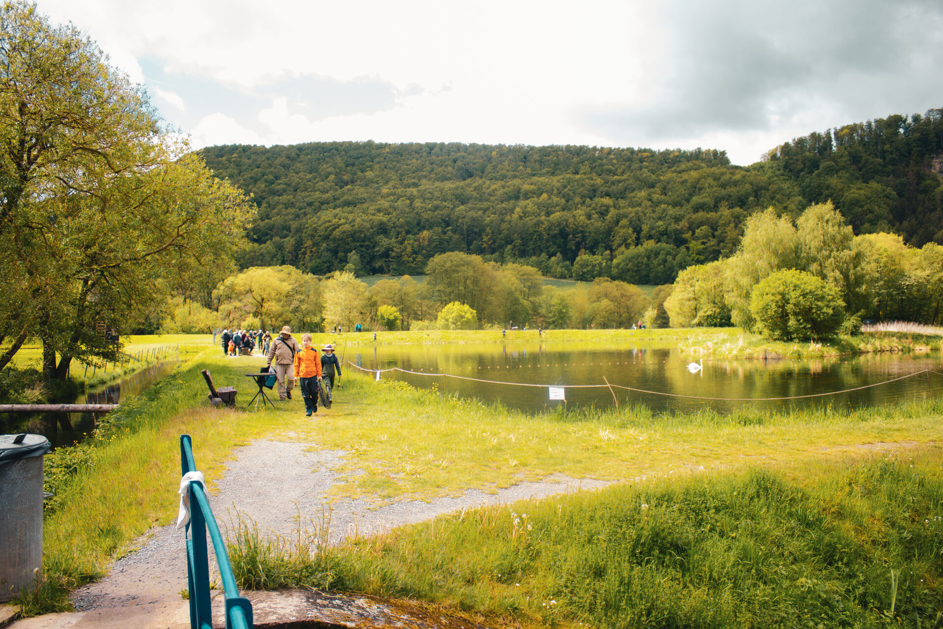 Forellenteich mit Uferweg und Geländer, dahinter bewaldete Hänge